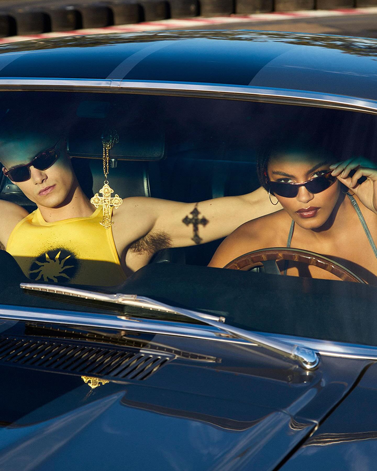 Two models in vintage car: man in yellow tank top with sun, woman in denim, both wearing sunglasses inside a blue classic car.