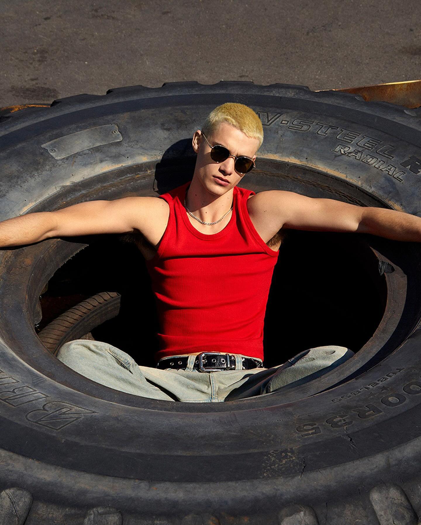 Male model in a red tank top poses inside a large tire outdoors, wearing round sunglasses and light wash jeans with a black belt.