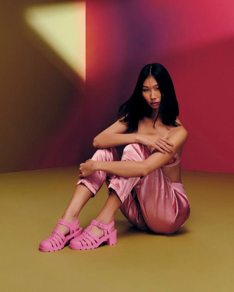 Young Asian woman posing seated in pink satin outfit and pink heeled sandals in a colorful photo studio setting. Fashion shot.