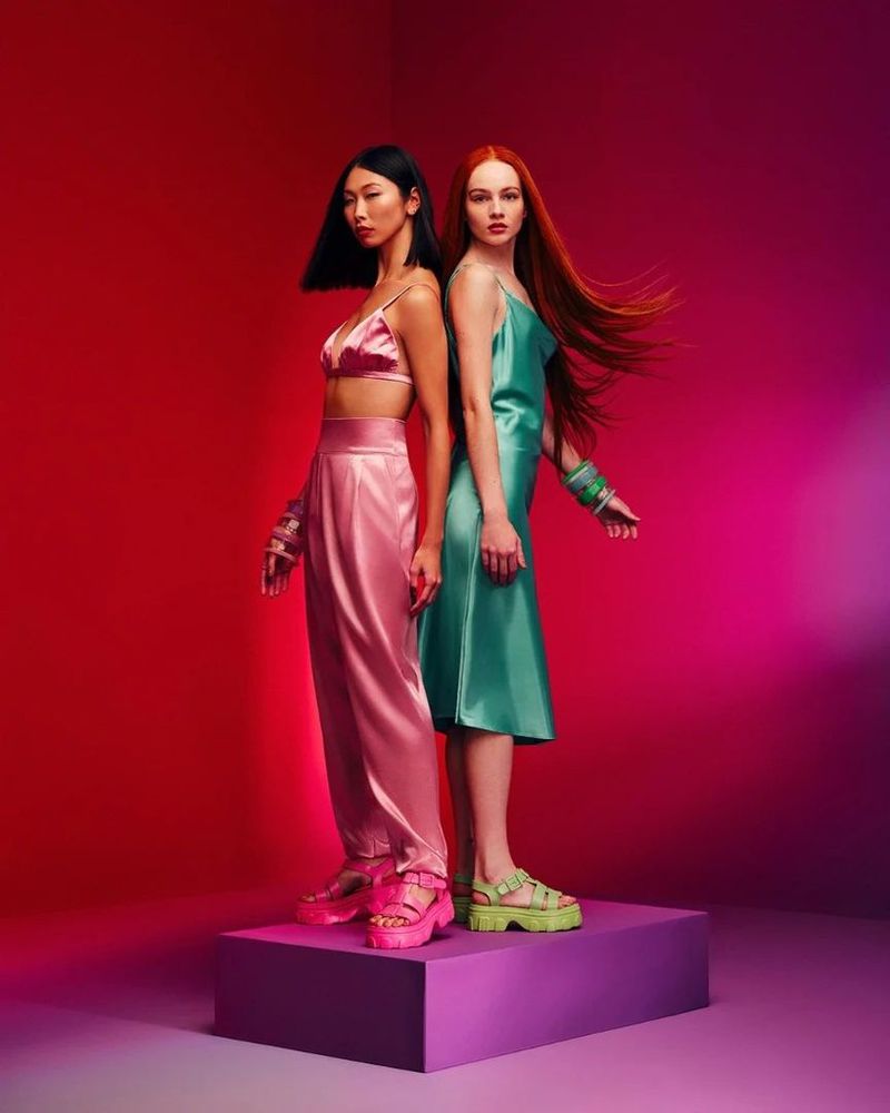 Two fashion models stand back-to-back on a platform wearing matching outfits and platform sandals against a vibrant red and purple backdrop.