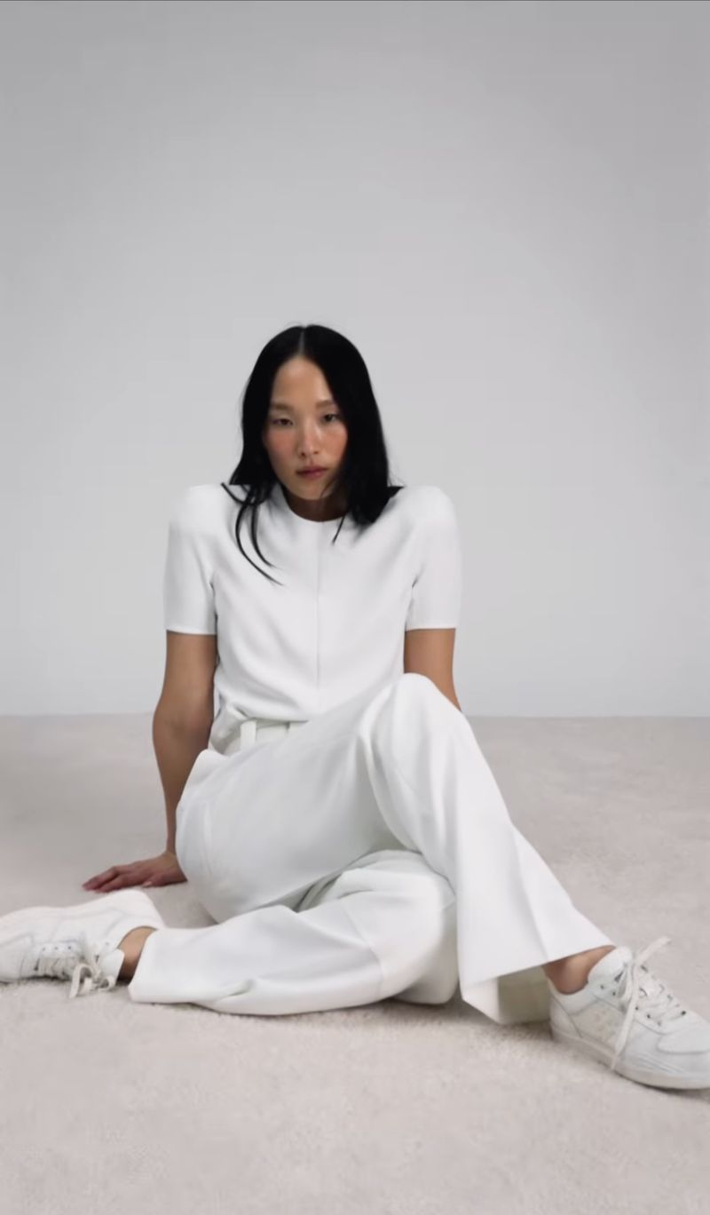 Adult female fashion model in white t-shirt and pants sitting on white carpet in studio. Minimalist aesthetic and neutral color palette.