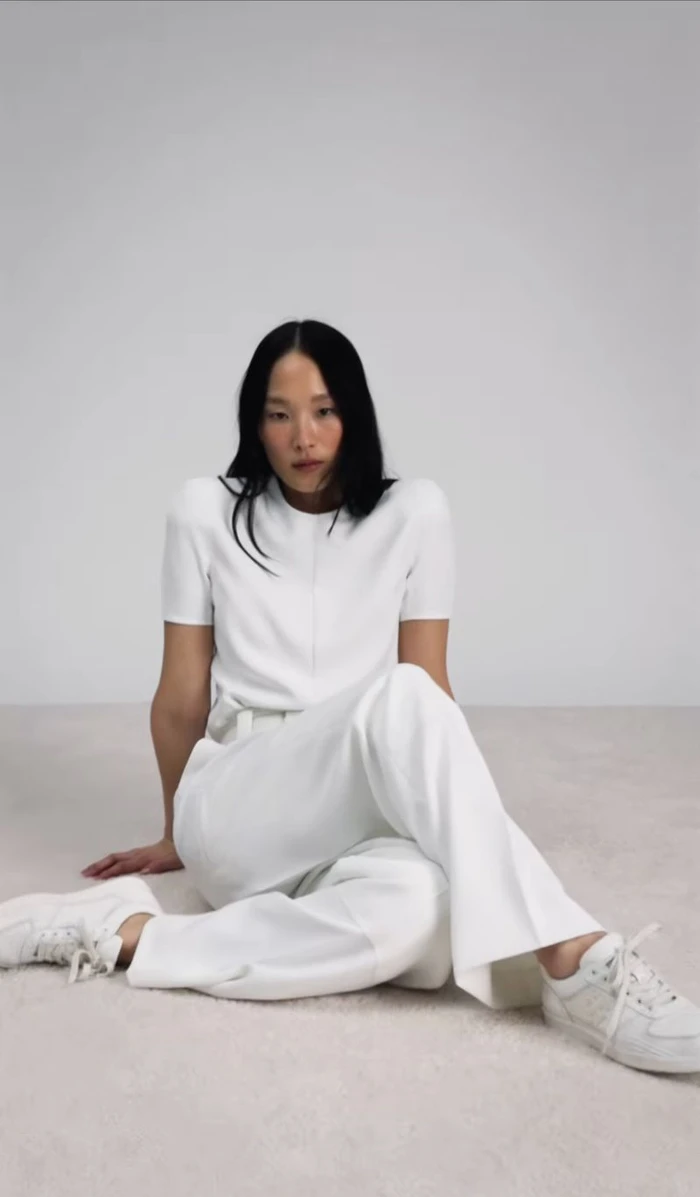 Model Yoonmi Sun seated on a grey seamless backdrop wearing a white t-shirt, white trousers, and white sneakers.
