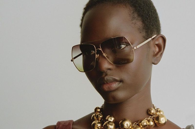 Close-up of a dark-skinned woman wearing gold-framed sunglasses and a gold beaded necklace against a pale neutral background.