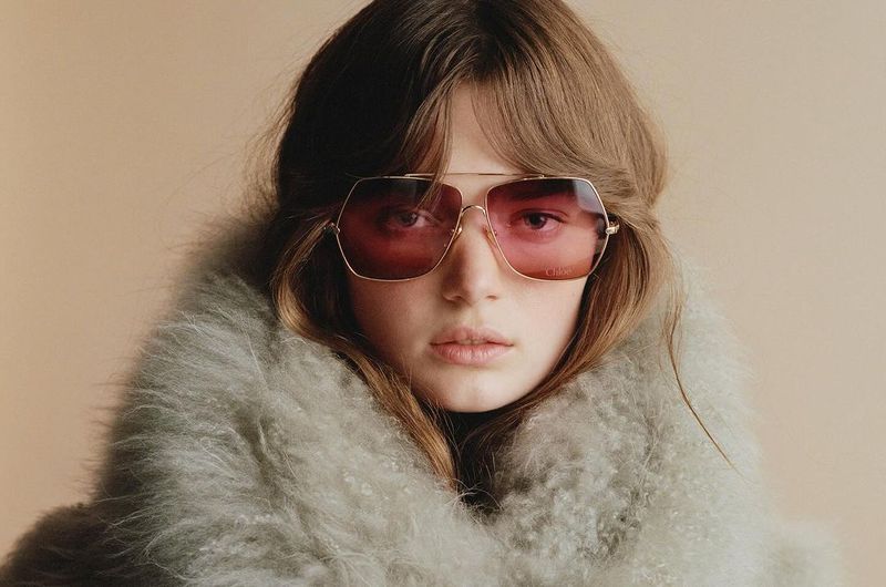 Close-up shot of a blonde woman with fair skin wearing oversized, gradient, aviator-style sunglasses and a grey fur coat.