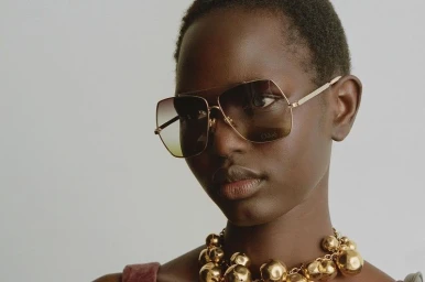 Close-up portrait of a model wearing oversized aviator sunglasses and a chunky gold sphere statement necklace.