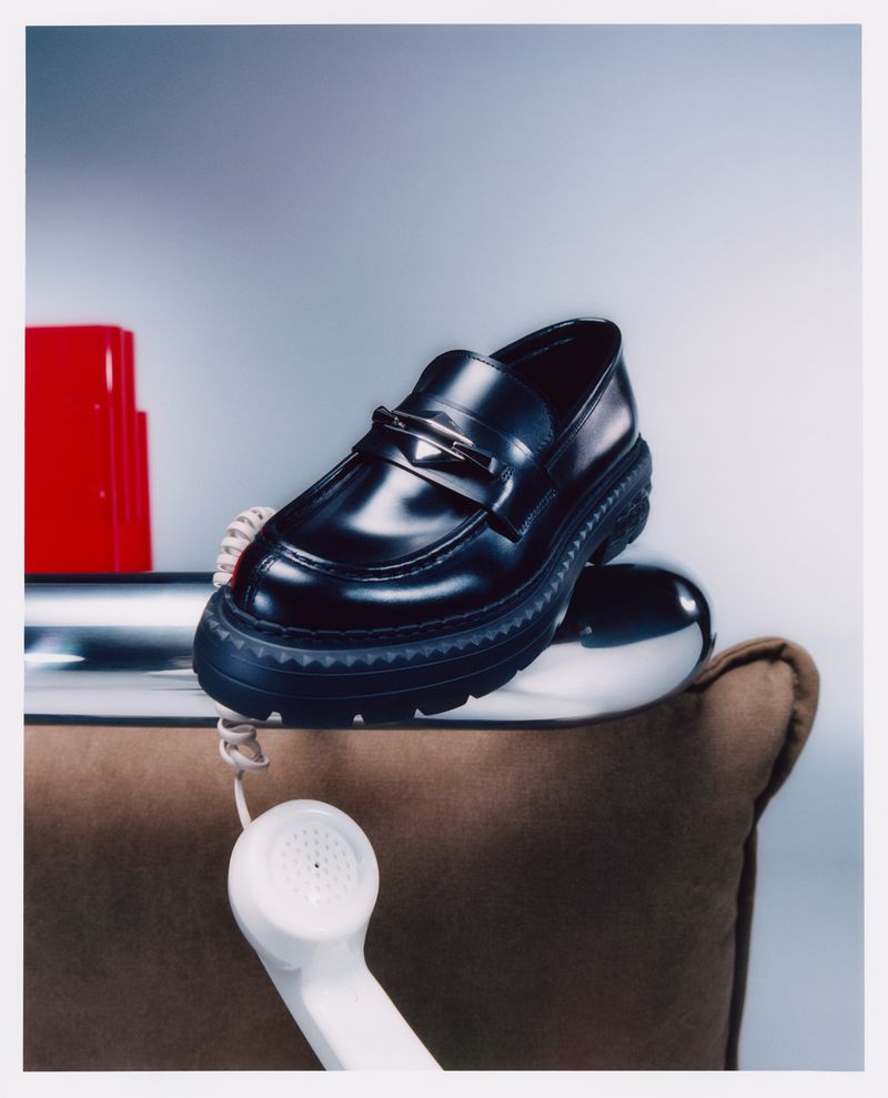Studio shot of a black leather loafer shoe balanced on a reflective silver dome next to a vintage rotary telephone and red object.