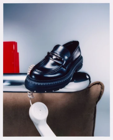 Studio shot of a black leather loafer shoe balanced on a reflective silver dome next to a vintage rotary telephone and red object.