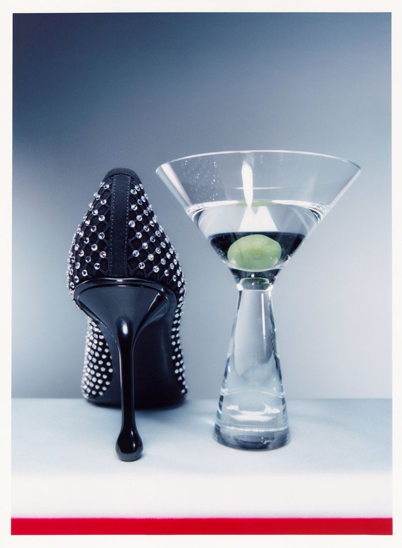 High heel shoe embellished with rhinestones positioned beside a clear martini glass with an olive, set against a gradient backdrop.