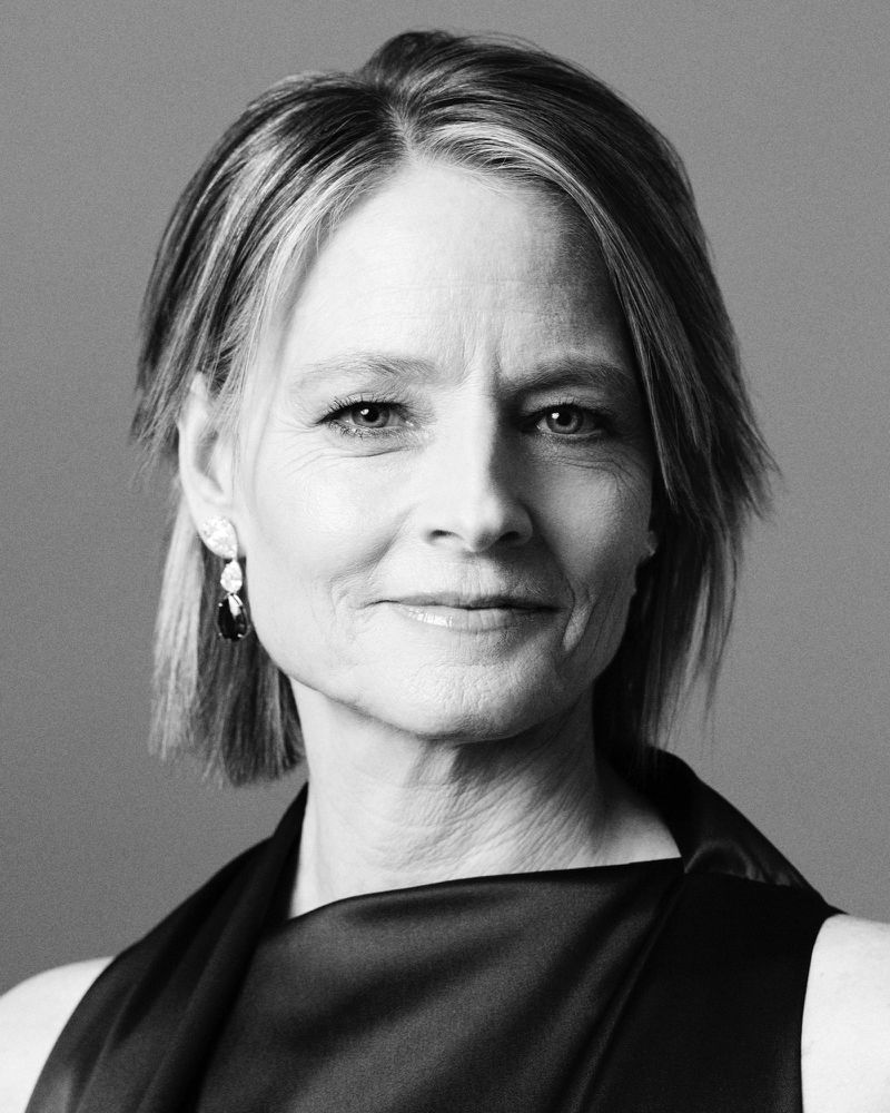 Black and white portrait of an adult woman with short hair, wearing a sleeveless satin top and teardrop earrings, against a neutral background.