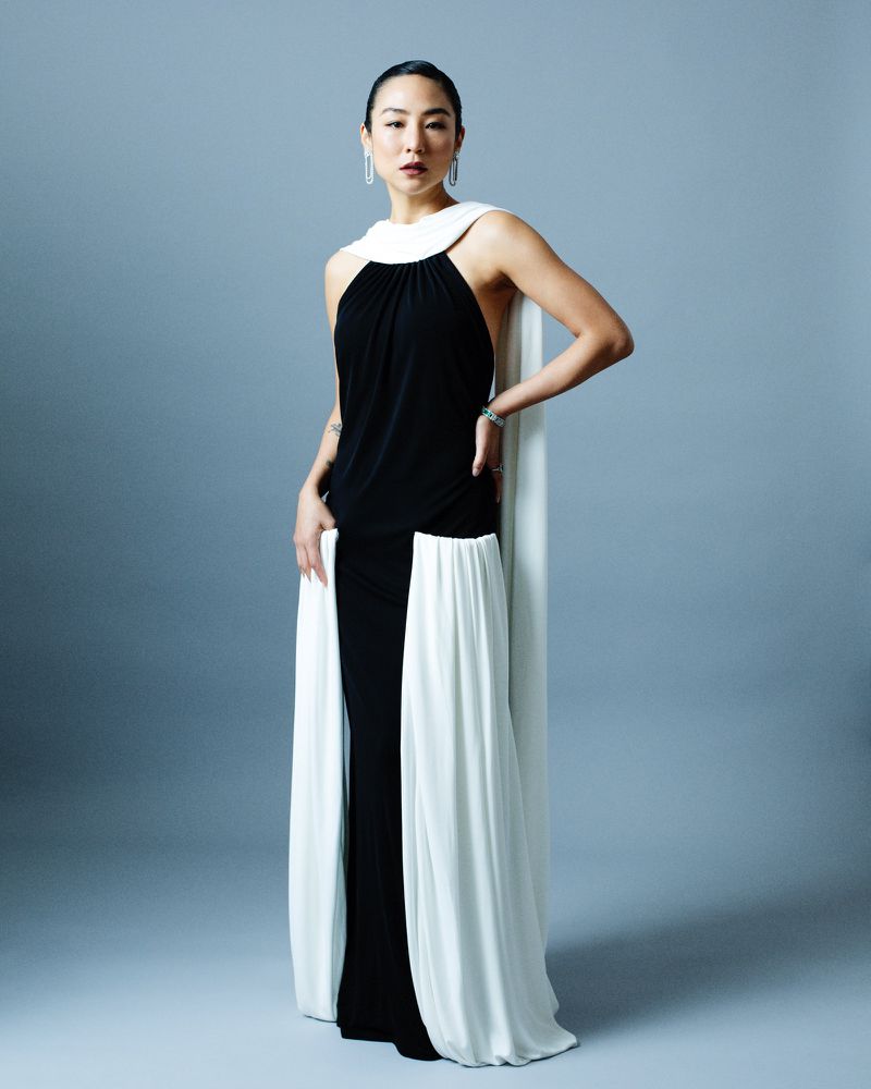 Elegant woman in black and white gown posing in studio with neutral backdrop; floor-length dress, sleeveless design, and statement earrings.