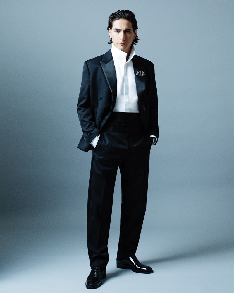 Full-length shot of an adult male model in a black tuxedo and patent leather shoes, hands in pockets, posing against grey backdrop.