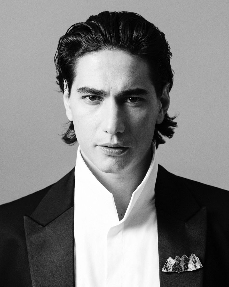 Headshot of a handsome adult male model with dark hair wearing a black tuxedo jacket and white shirt against a gray background.
