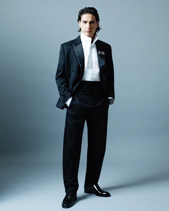 Man wearing a black tuxedo and white collared shirt standing against a grey studio backdrop