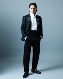 Man wearing a black tuxedo and white collared shirt standing against a grey studio backdrop