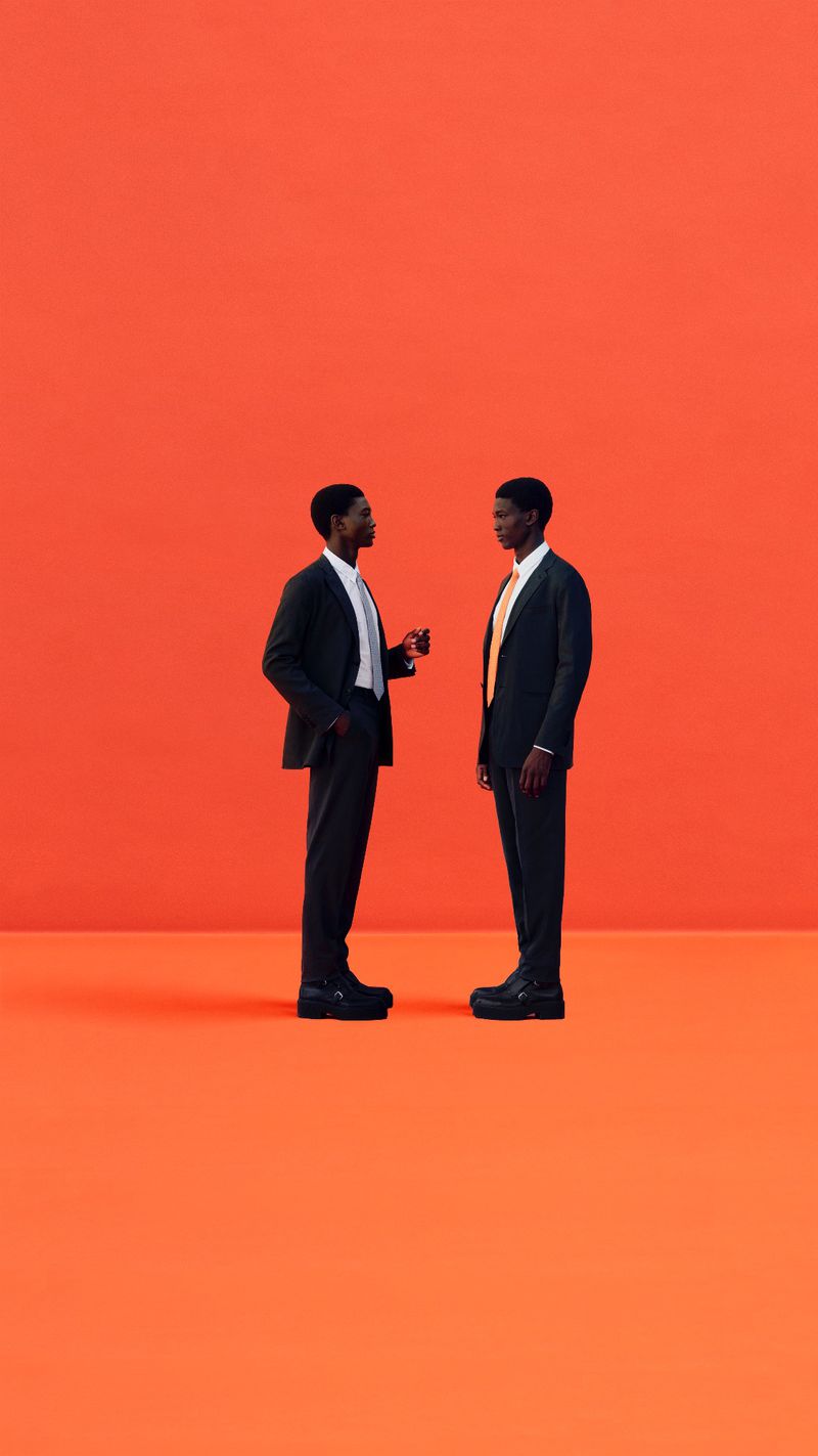 Two Black men in suits standing face to face on an orange studio backdrop, professionally lit for business or fashion editorial.