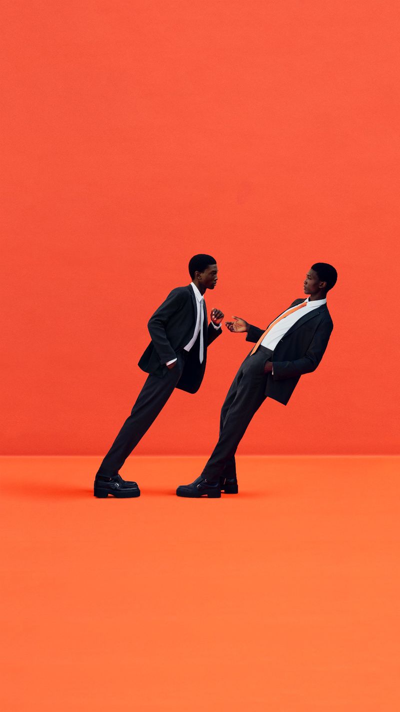 Two Black male models in suits pose against a studio background of orange, showcasing modern tailoring and minimalist fashion.