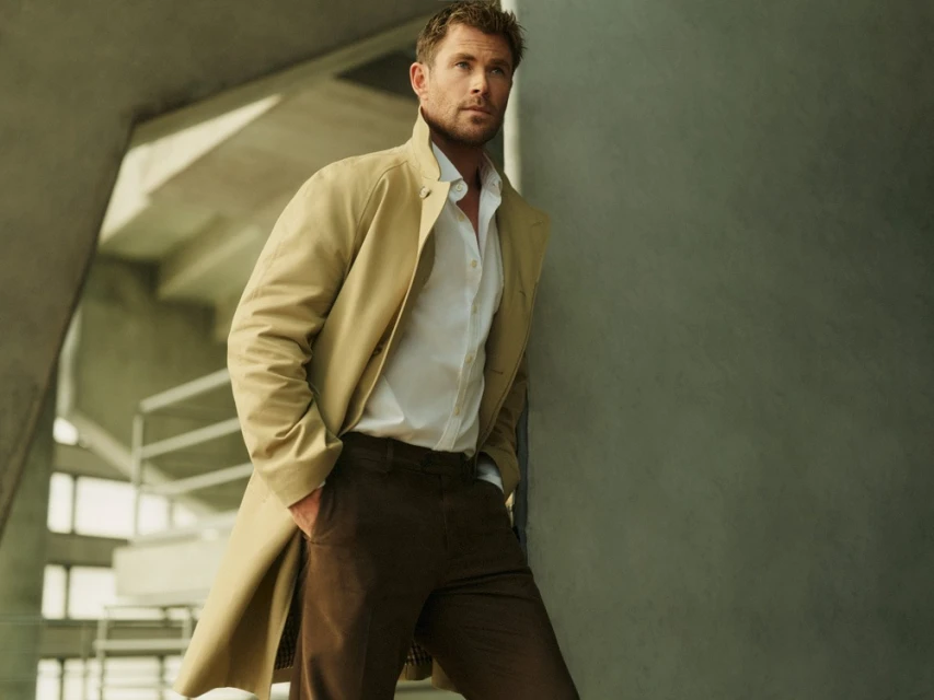Portrait of Chris Hemsworth in a trench coat leaning against a wall in an urban setting with natural light, wearing an unbuttoned white shirt.