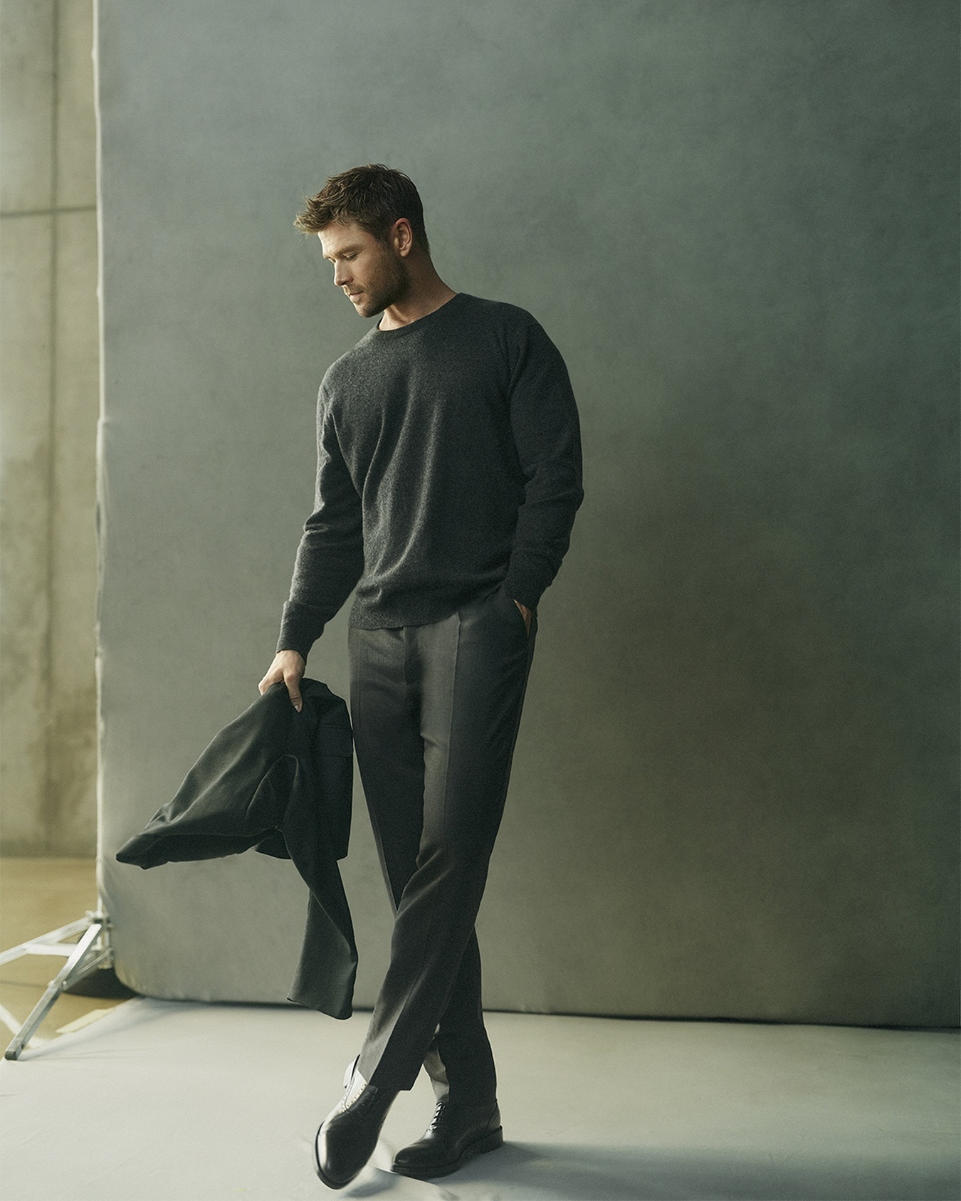 Chris Hemsworth standing in dark sweater and trousers holding a jacket against a grey studio backdrop