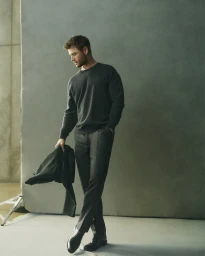 Chris Hemsworth standing in dark sweater and trousers holding a jacket against a grey studio backdrop