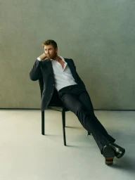 Chris Hemsworth seated on a black chair wearing a dark suit and white shirt against a neutral grey studio backdrop