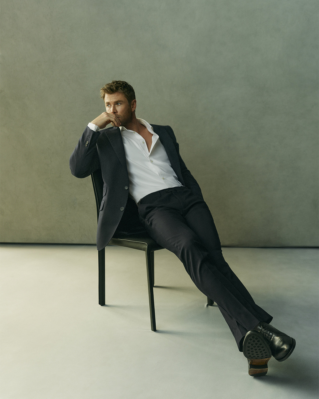 Chris Hemsworth in a dark suit and white shirt seated on a black chair against a grey studio backdrop.