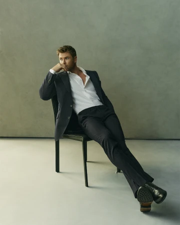 Chris Hemsworth in a dark suit and white shirt seated on a black chair against a grey studio backdrop.