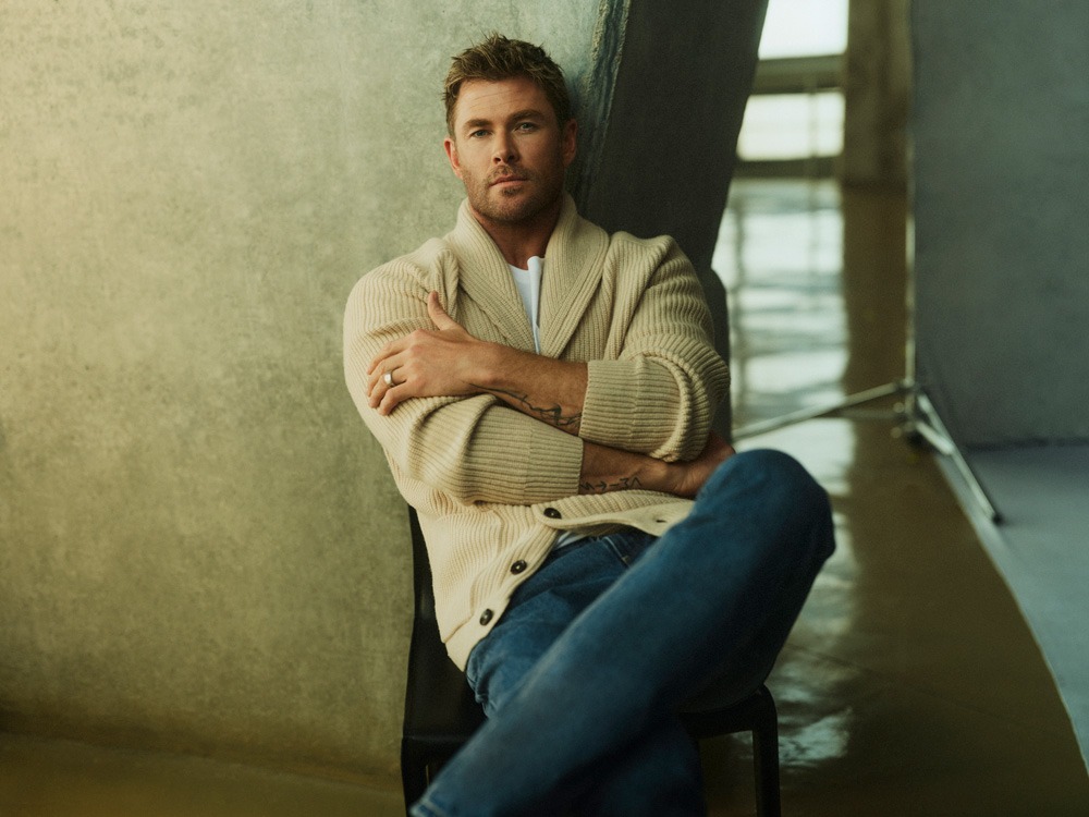 Portrait of Chris Hemsworth in a cream cardigan, blue jeans, posing indoors against a neutral background. Professional celebrity shot.