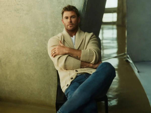 Portrait of Chris Hemsworth in a cream cardigan, blue jeans, posing indoors against a neutral background. Professional celebrity shot.