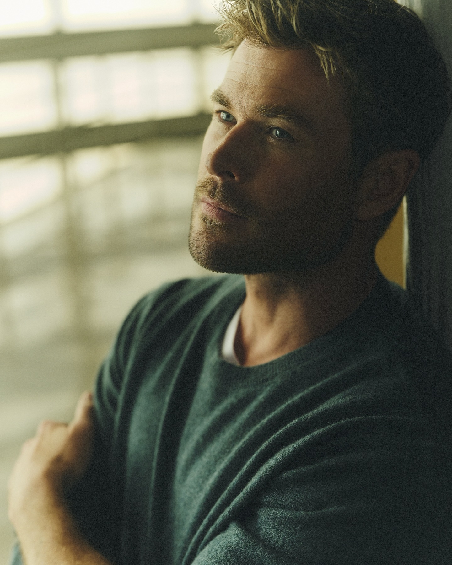 Portrait of a handsome adult man with fair skin and stubble. He has dark blond hair and is wearing a green sweater indoors.
