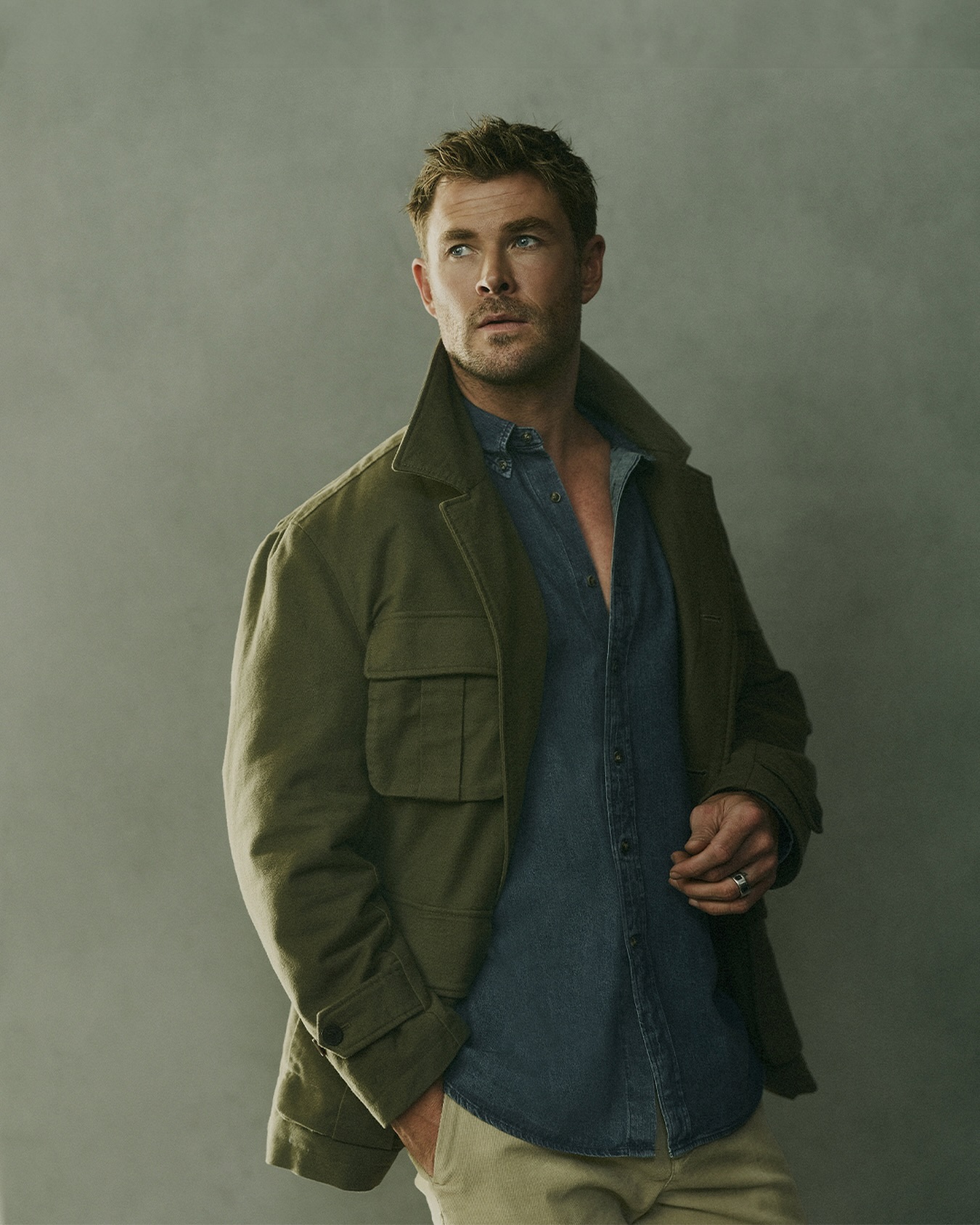 Portrait of Chris Hemsworth looking left. He wears an olive jacket over a denim shirt, paired with beige pants against a neutral backdrop.
