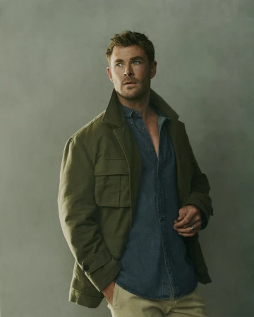 Portrait of Chris Hemsworth looking left. He wears an olive jacket over a denim shirt, paired with beige pants against a neutral backdrop.