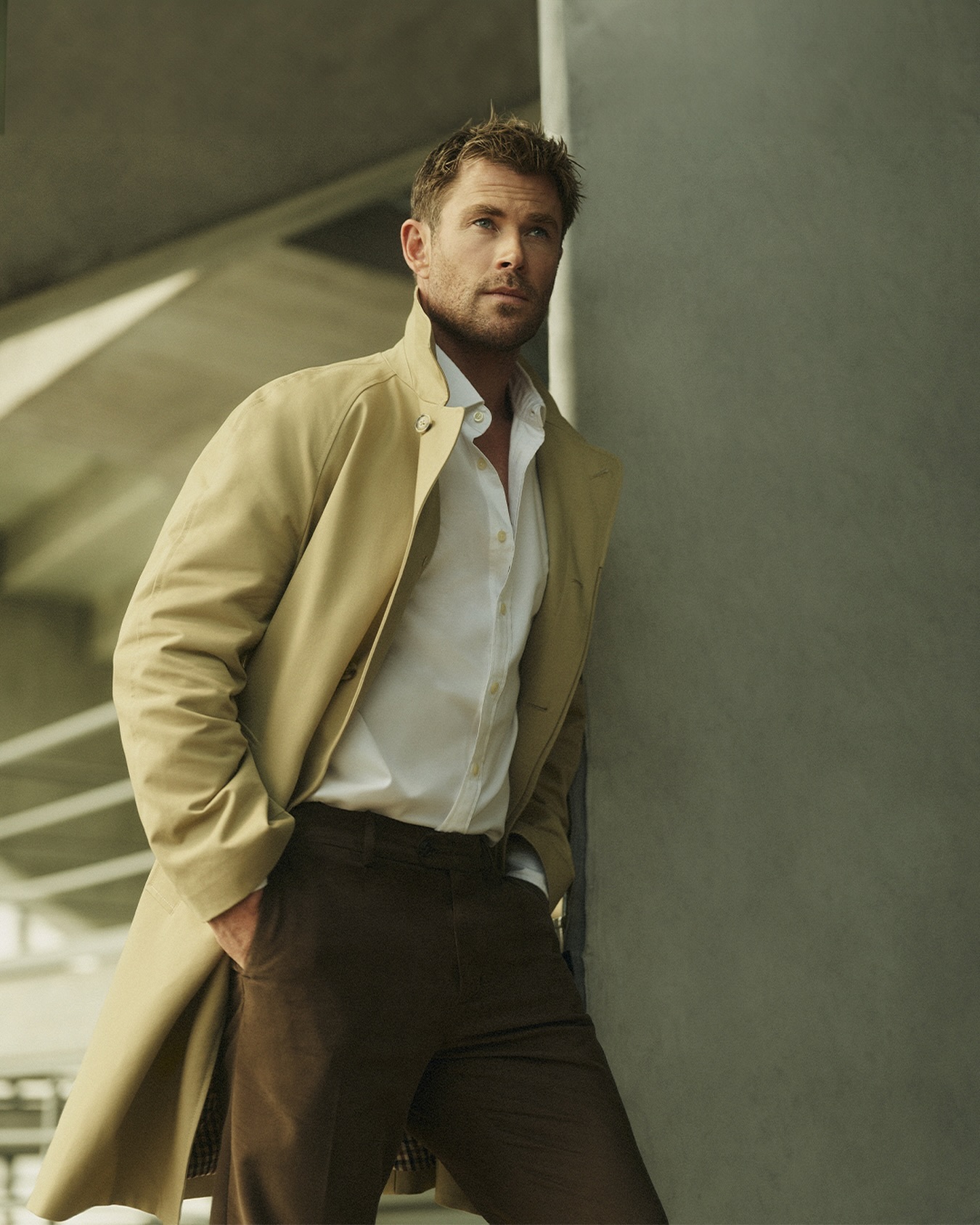 Portrait of a handsome adult male in a tan trench coat, white shirt, and brown pants leaning against a gray wall indoors.