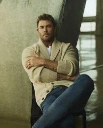 Chris Hemsworth wearing a cream cardigan and blue jeans, seated against a textured concrete pillar.