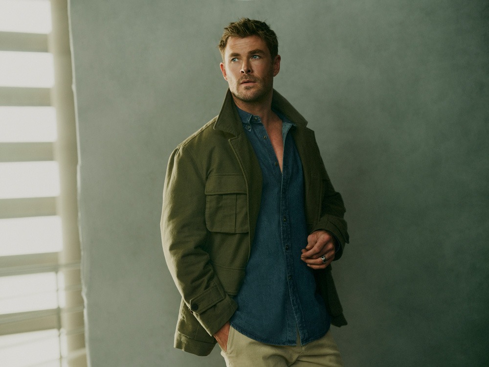 Attractive adult male poses indoors near window with horizontal blinds, wearing an olive-green jacket, button-down shirt, and tan pants.