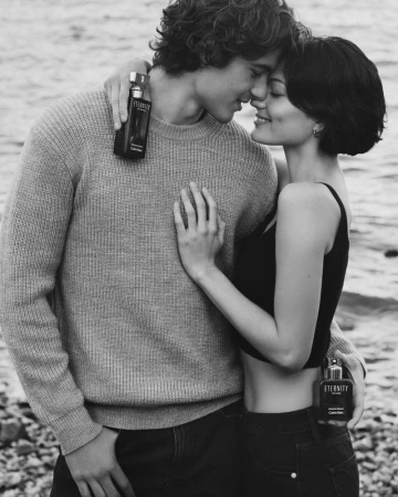 Black and white portrait of a romantic couple embracing by the water, holding a fragrance bottle against a backdrop of water and rocks.