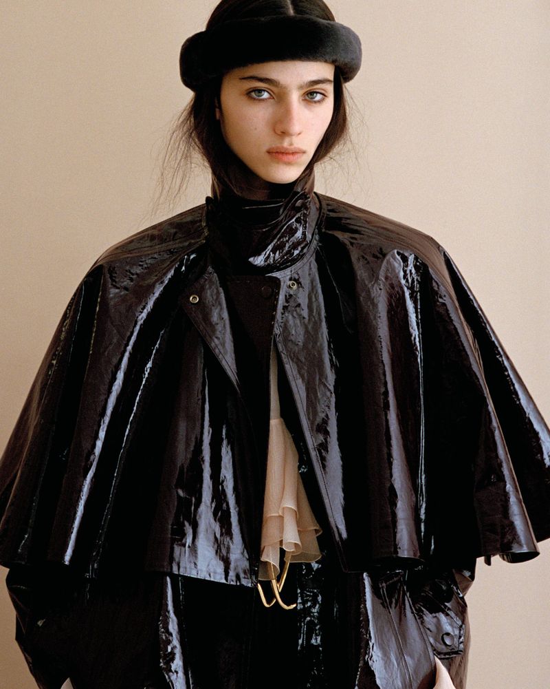 Portrait of a model wearing a shiny black cape and a fur hat against a neutral backdrop in a fashion photography style.
