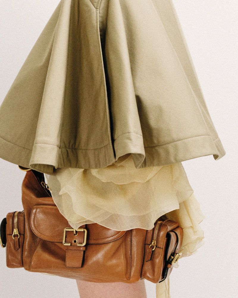 A brown leather handbag is accessorized with a light yellow skirt and khaki jacket in front of a solid neutral background.