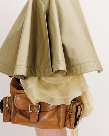 A brown leather handbag is accessorized with a light yellow skirt and khaki jacket in front of a solid neutral background.