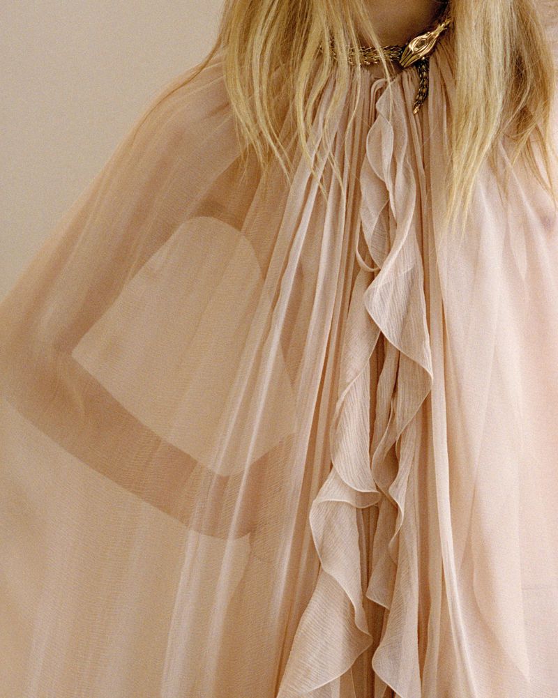 Woman wearing a sheer, flowing pink gown with ruffles and a snake necklace, captured in a close-up shot, highlighting the dress's texture.