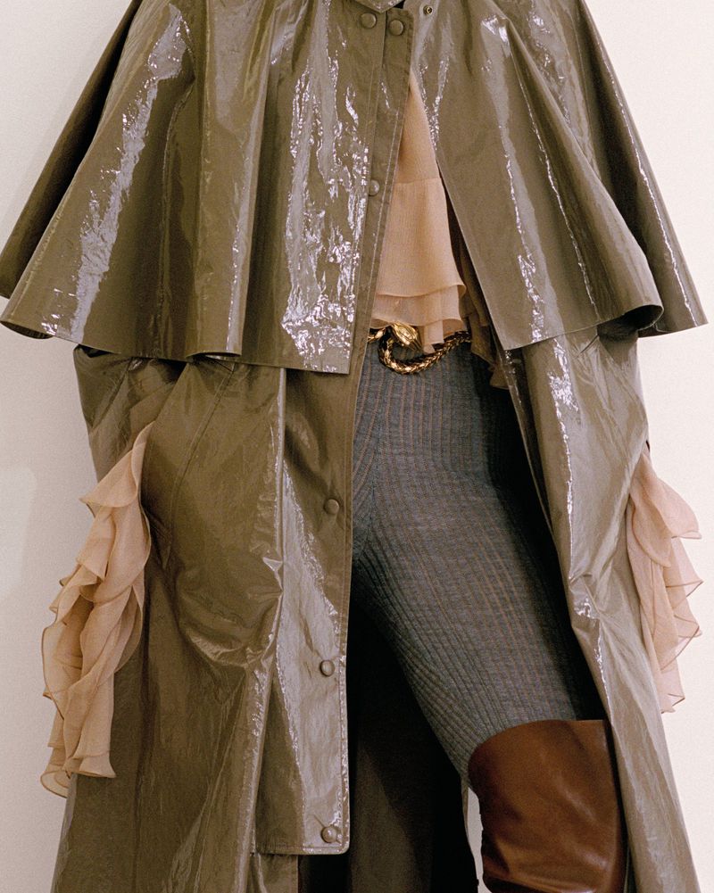 Model showcases shiny trench coat with cape detail, paired with ribbed leggings, brown boots, and a snake belt buckle against white.