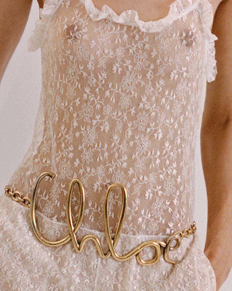 Woman in a white lace dress with a gold 'Chloe' belt standing in front of a white background in a close up shot, fashion photography.