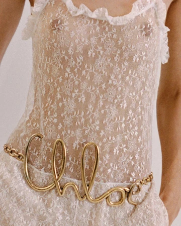 Woman in a white lace dress with a gold 'Chloe' belt standing in front of a white background in a close up shot, fashion photography.