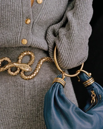 Close-up of a woman's torso wearing a grey sweater and a gold snake belt, accessorized with a blue leather purse. Fashion shot.