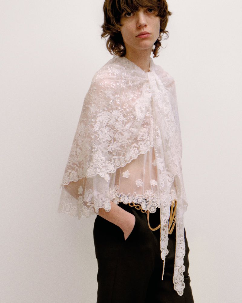 Woman in a white lace cape with black pants and a gold chain belt poses against a neutral background in a fashion shot.