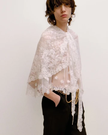 Woman in a white lace cape with black pants and a gold chain belt poses against a neutral background in a fashion shot.