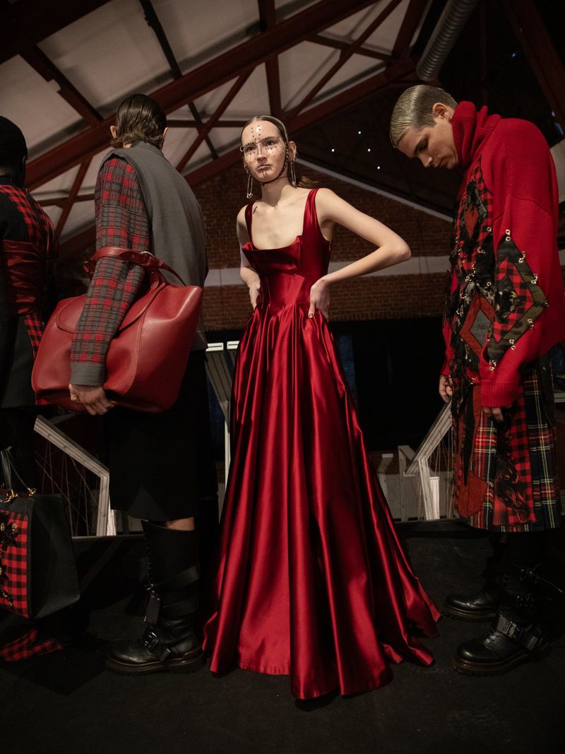 A runway fashion show featuring a woman in a red gown. Other models stand on the sides wearing red and black tartan outfits.