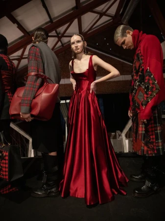 A runway fashion show featuring a woman in a red gown. Other models stand on the sides wearing red and black tartan outfits.