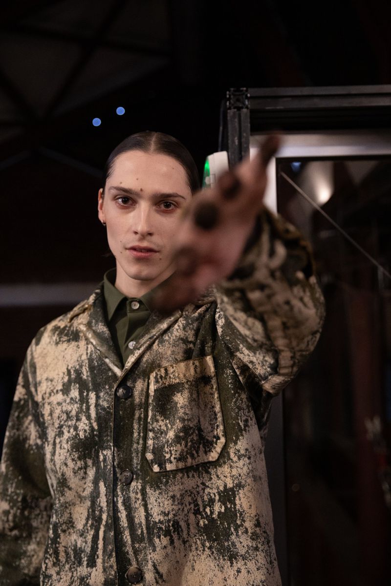 Portrait of a young man wearing a patterned jacket, hand extended toward camera in a dimly lit indoor setting.