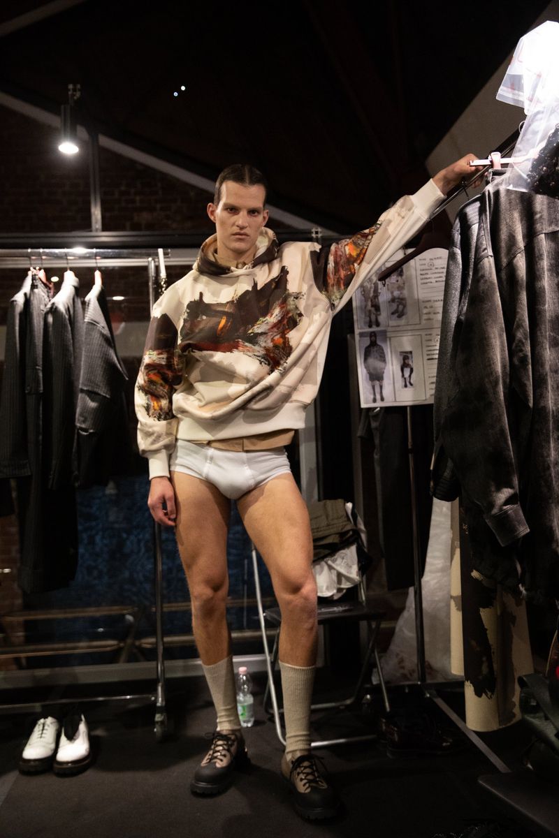 Male fashion model posing in underwear, socks, and boots while wearing a patterned hoodie in an indoor studio setting.