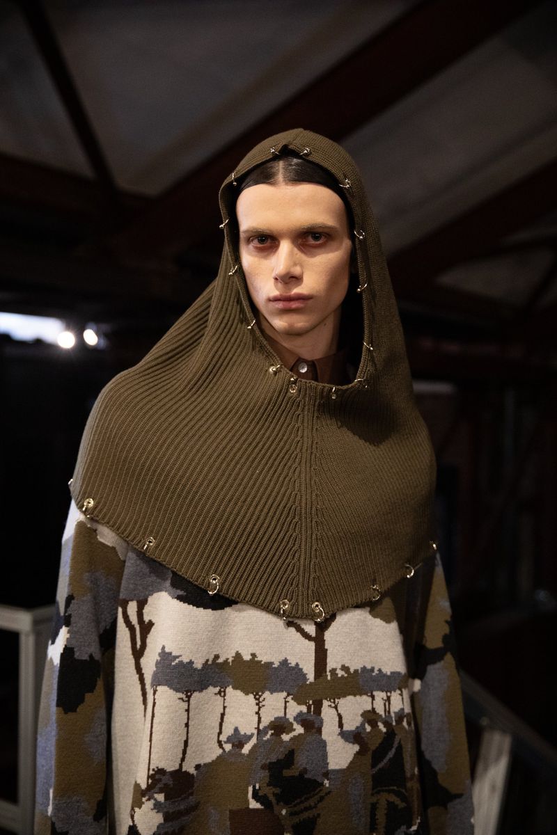 Close-up studio shot of a fashion model wearing an olive green hooded head covering and printed sweater with tree silhouettes.